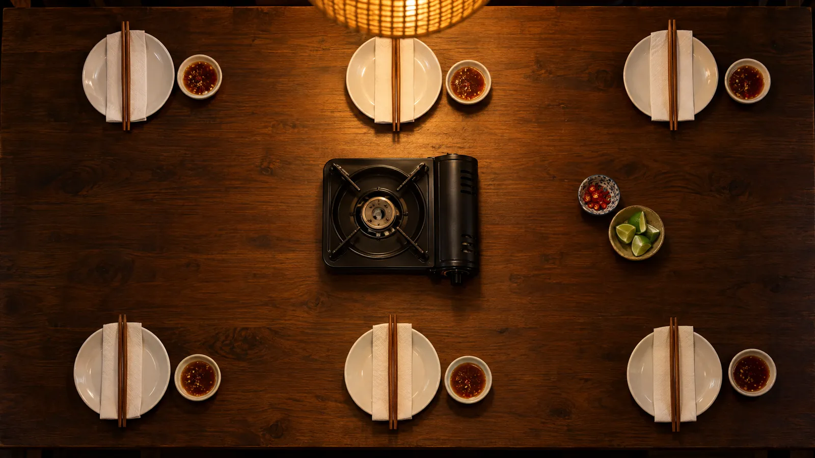 A dark wooden table set for six from above, plates, chopsticks and dipping sauces around a gas burner